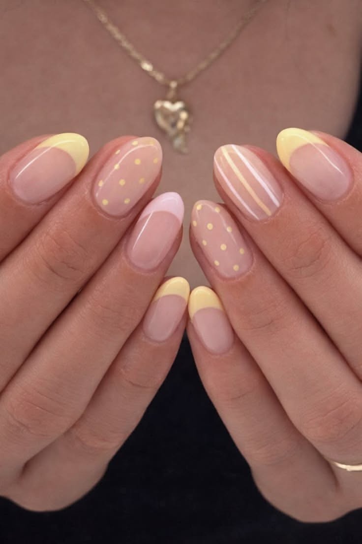 Close-up of hands with short, almond-shaped nails featuring a mix of pastel yellow and pink French tips, polka dots, and…