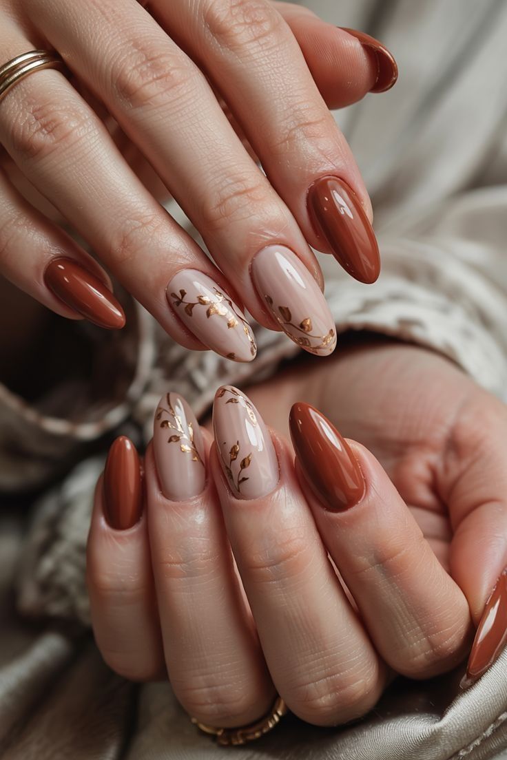 Close-up of manicured hands with long, almond-shaped nails. Alternating nails are solid reddish-brown or nude with gold leaf…