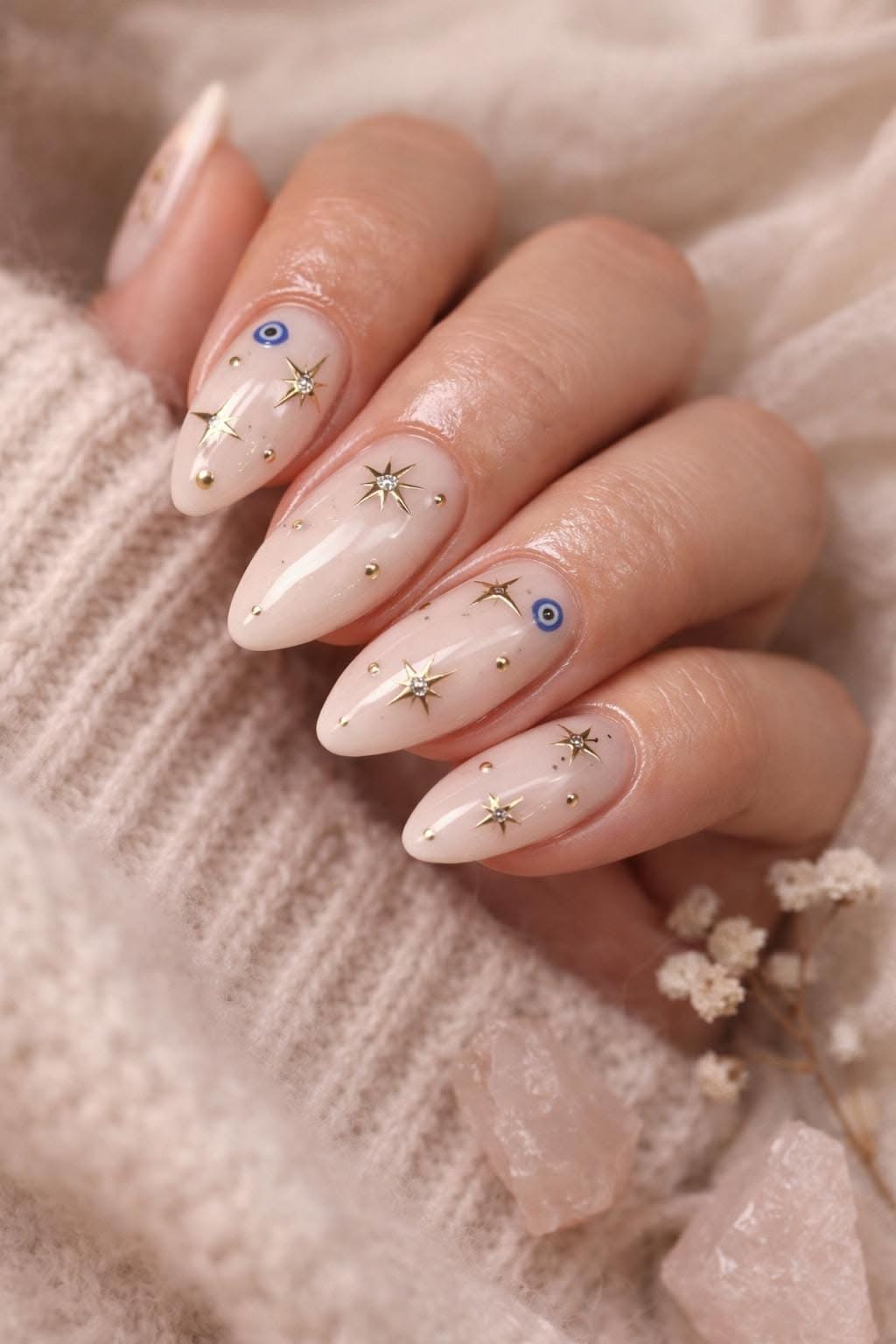 Almond-shaped nails with a nude base, gold star accents, small gold studs, and blue evil eye charms.