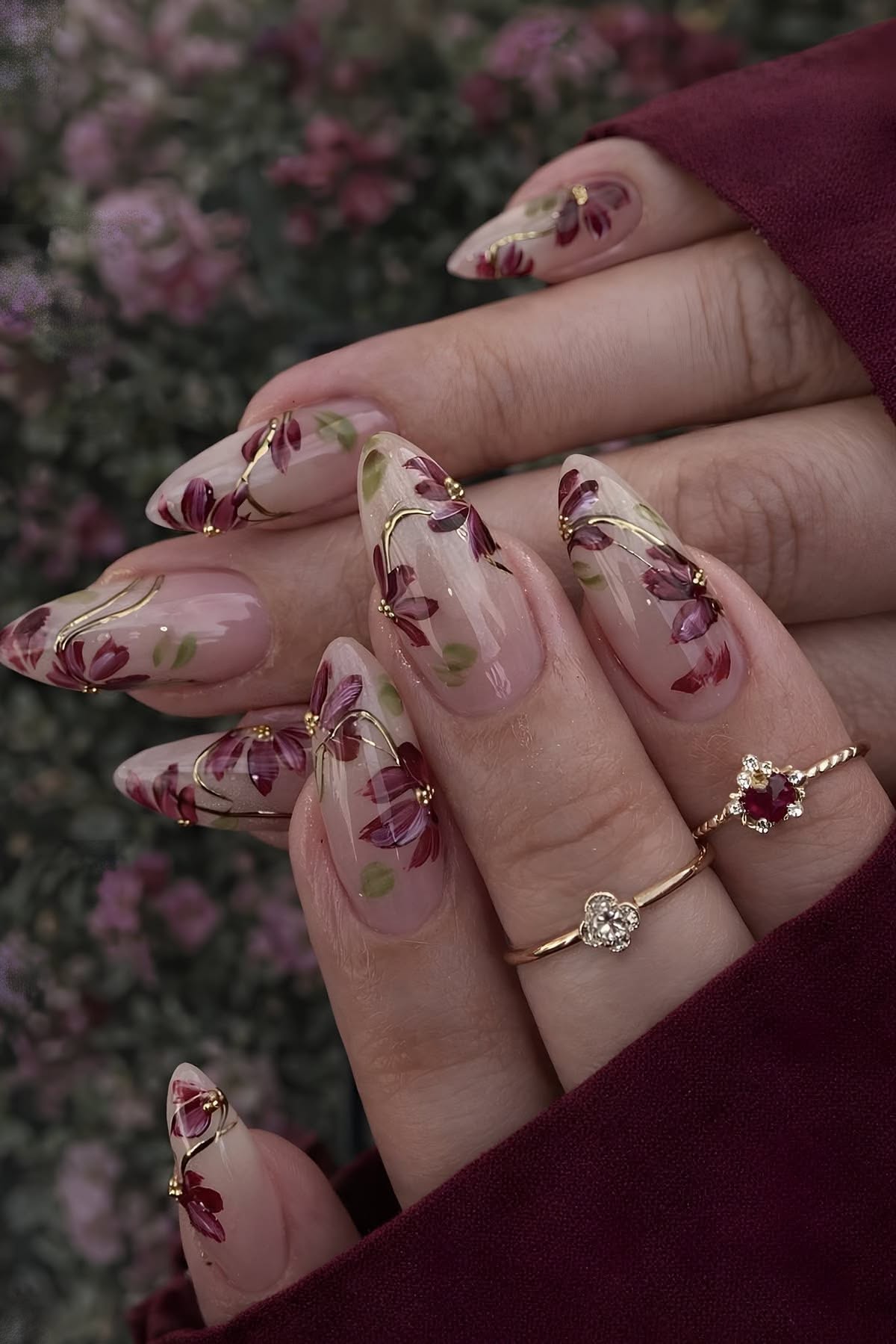 Close up of manicured hands with almond-shaped nails featuring floral designs in deep plum and gold accents, adorned with…