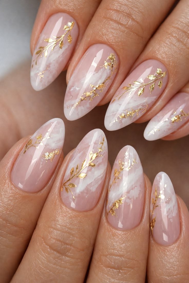 Almond-shaped nails with a translucent pink base, white marble-like designs, and scattered gold leaf in botanical patterns.