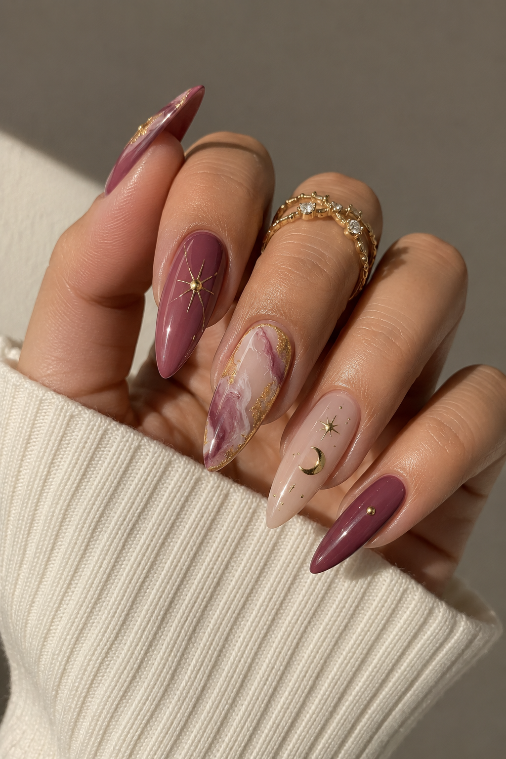 Almond-shaped nails in maroon, nude, and marble patterns with gold celestial decals.