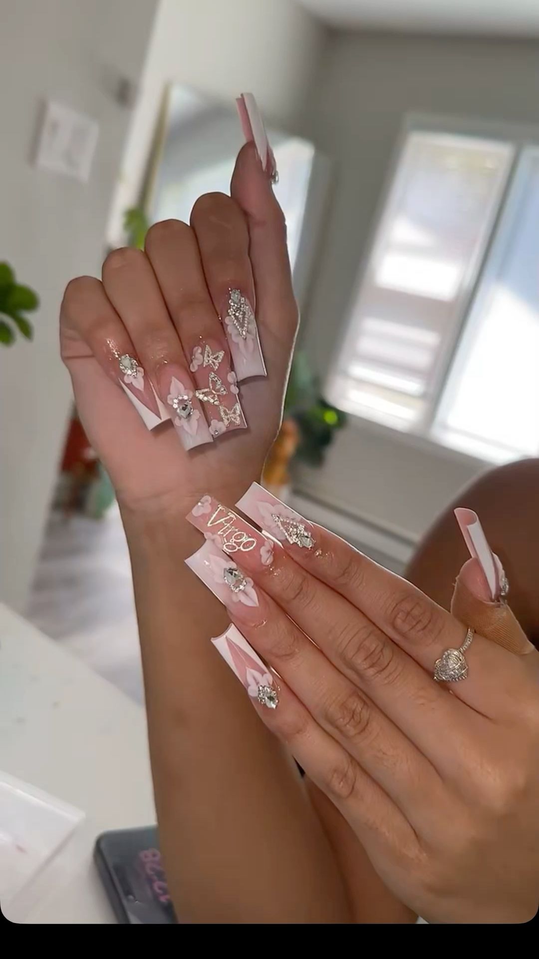 Close-up of a person's hands showing long, square-tipped freestyle nails with white and pink bases, adorned with rhinestones…