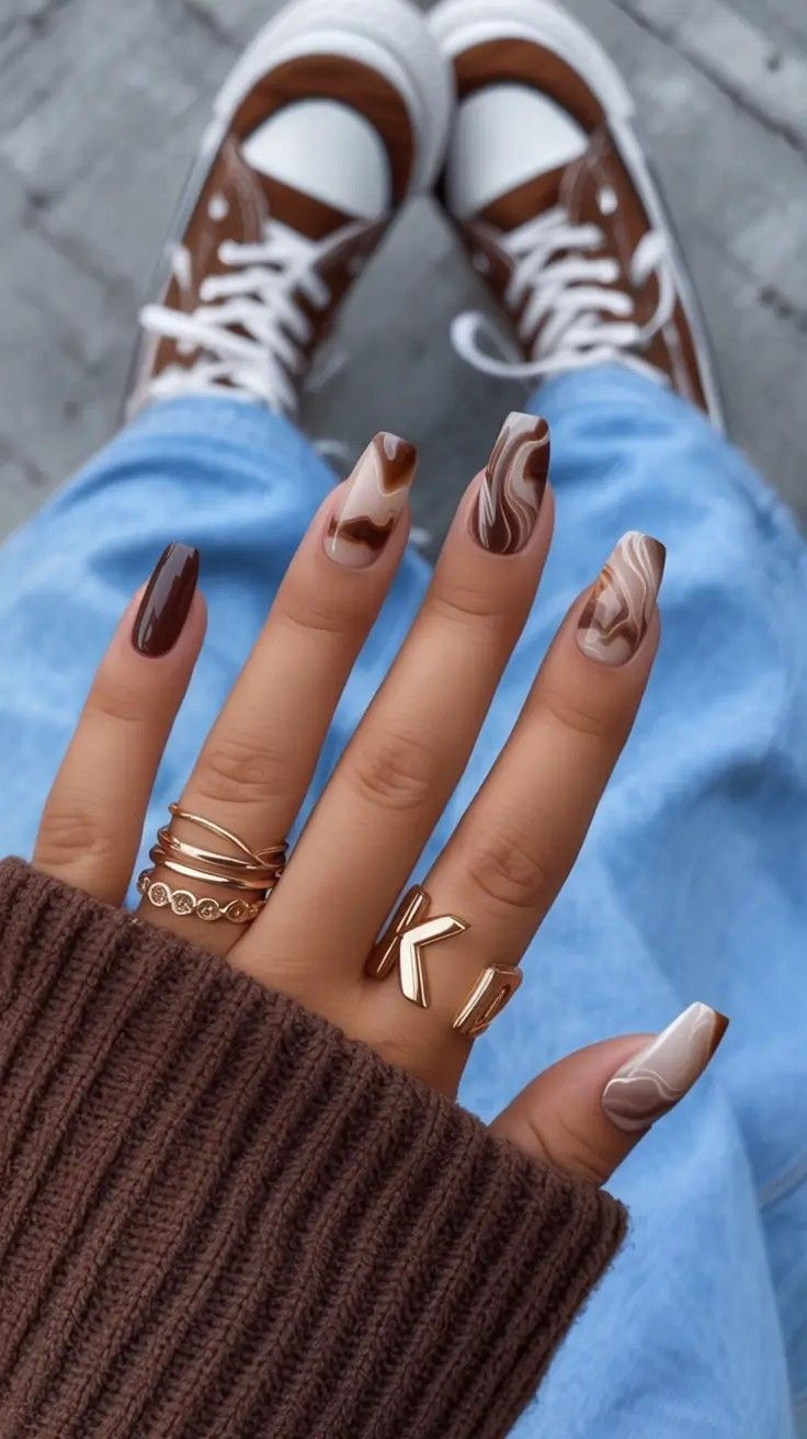 Close up of a hand with brown and beige marbled coffin nails, wearing gold rings and a brown sweater.