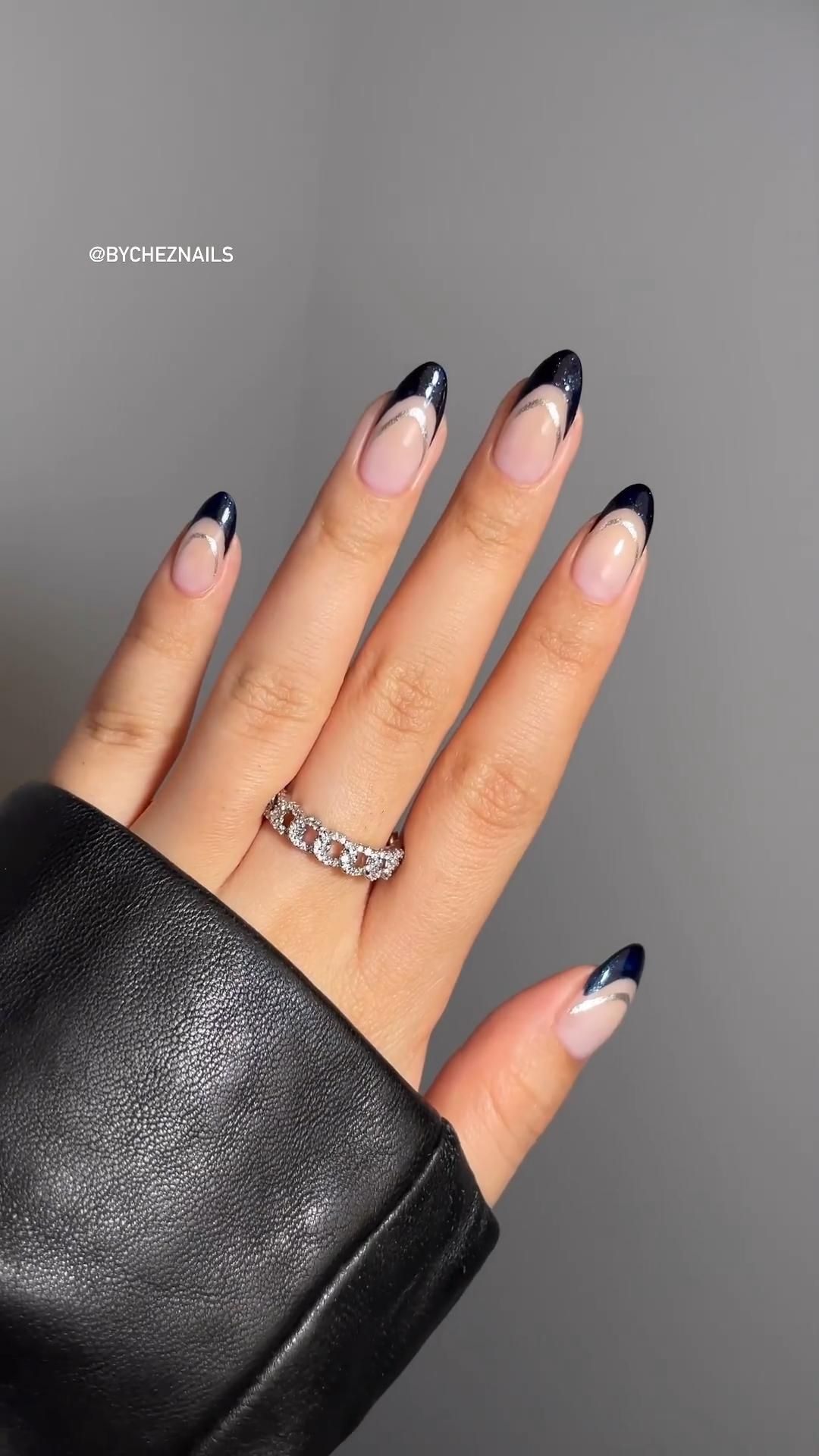 A hand displays nails with navy and silver glitter French tips, and a silver ring on the ring finger.