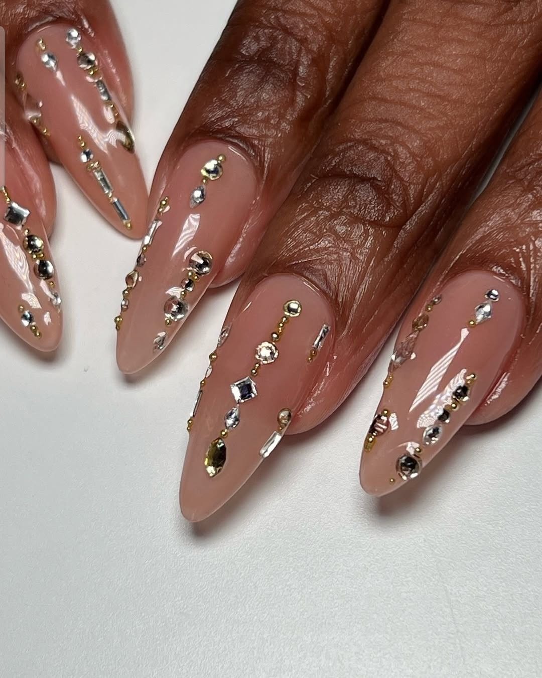 Pointed almond nails with a neutral base, adorned with rows of delicate gold and silver rhinestones, varying in size and…