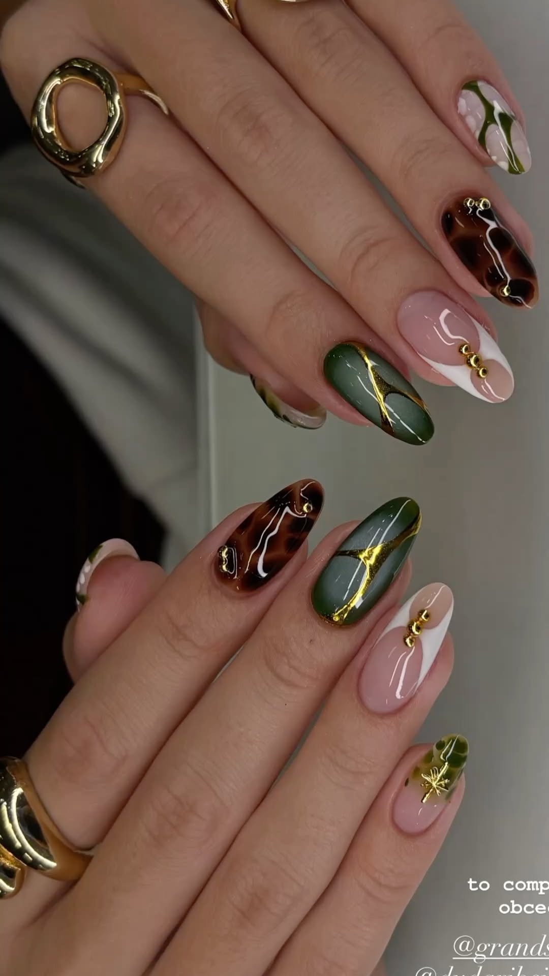 Close-up of hands with several almond-shaped nails featuring various designs in green, brown, and nude with gold accents and…