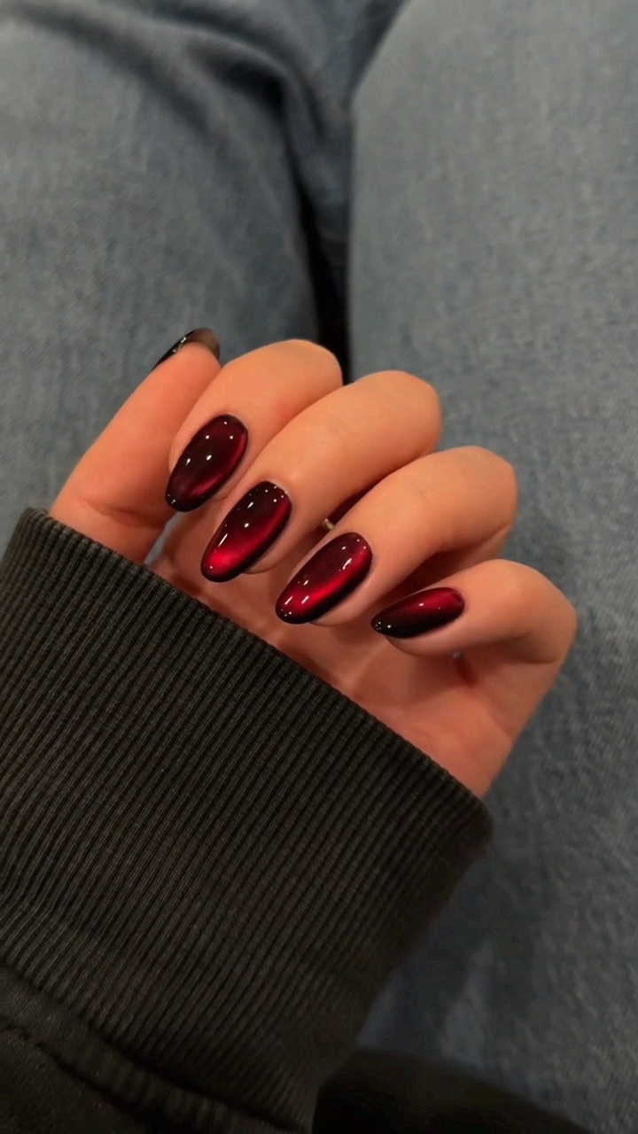 Hand with dark red cat eye nails, short almond shape, sleeve of a black ribbed sweater visible.