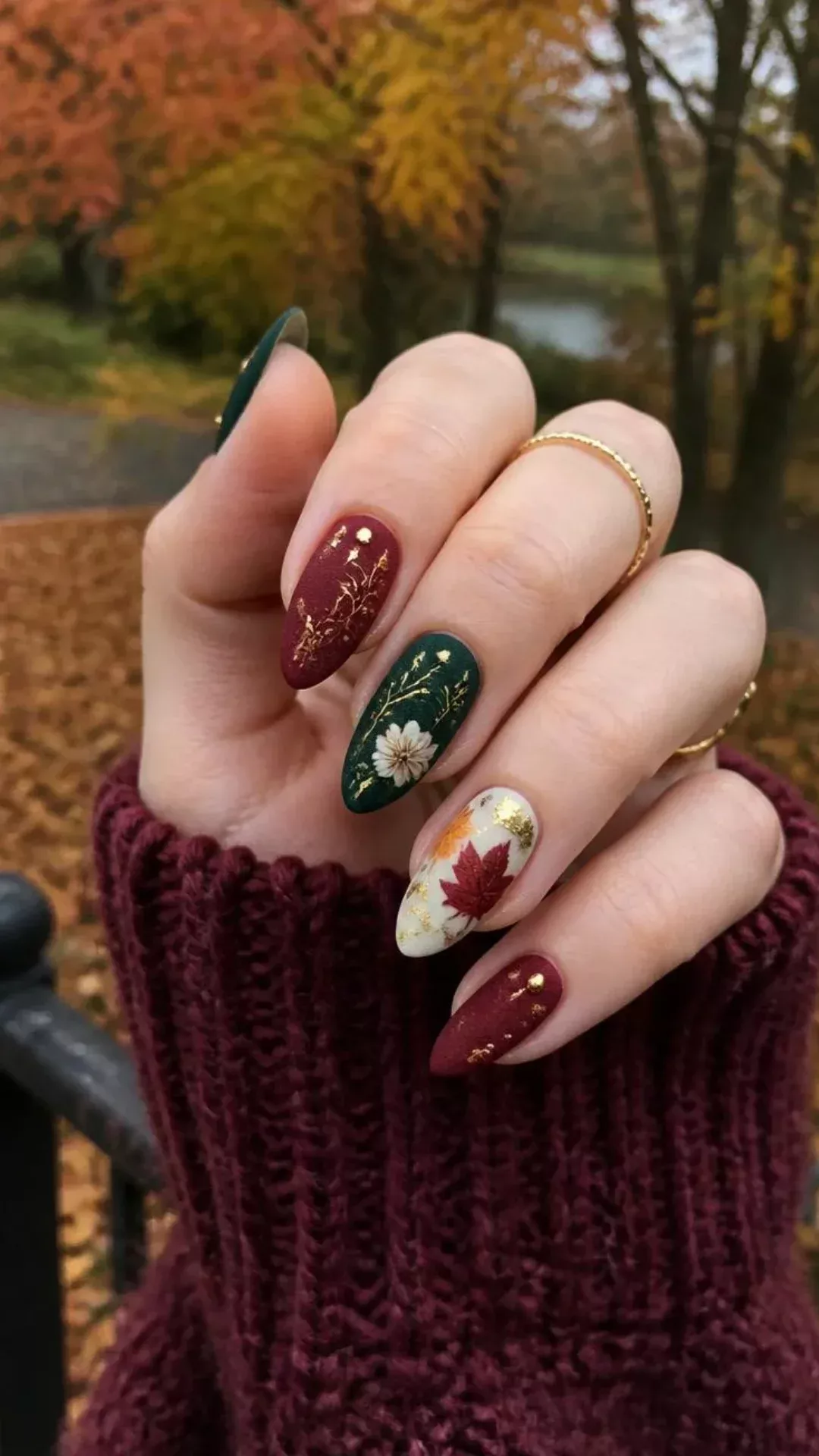 Close-up of a hand with almond-shaped fall-themed nails, featuring deep red, forest green, and ivory polishes with gold…