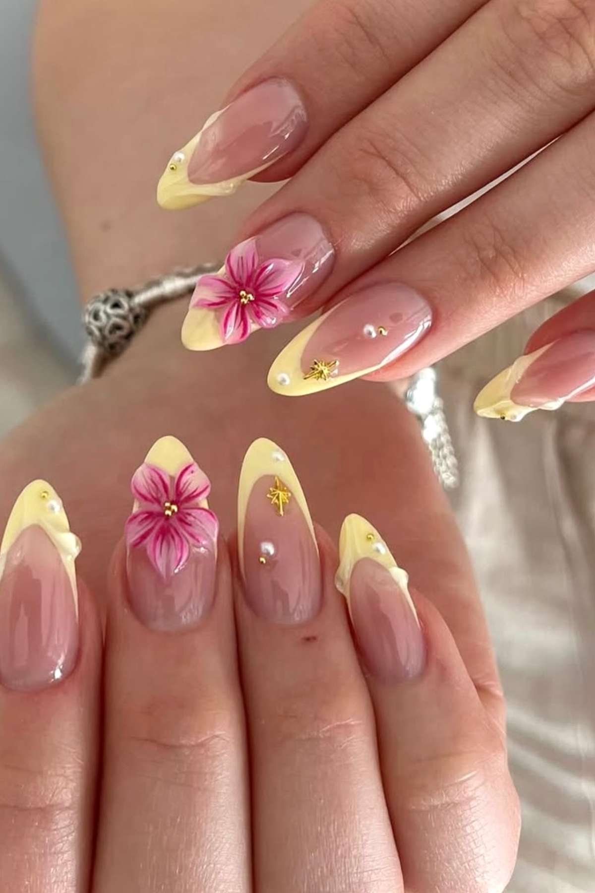 Close-up of a hand with almond-shaped nails featuring yellow tips, 3D pink flowers, gold stars, and small pearl…