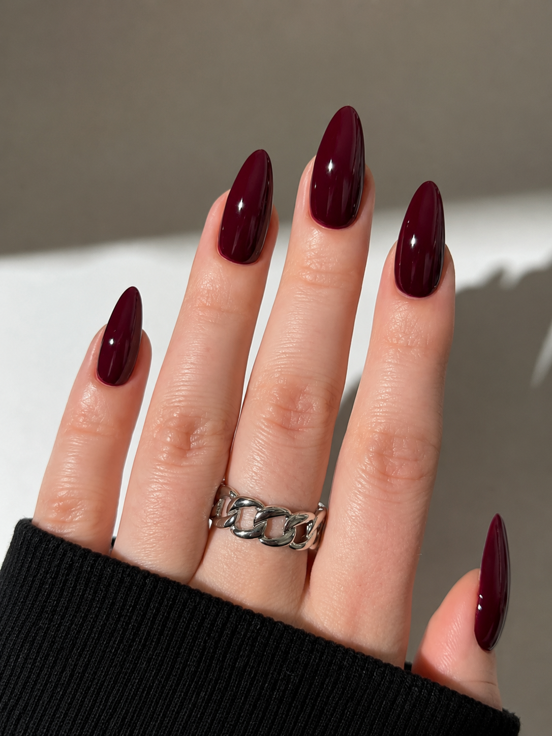 Hand with long, glossy burgundy almond-shaped nails, wearing a silver chain-link ring.