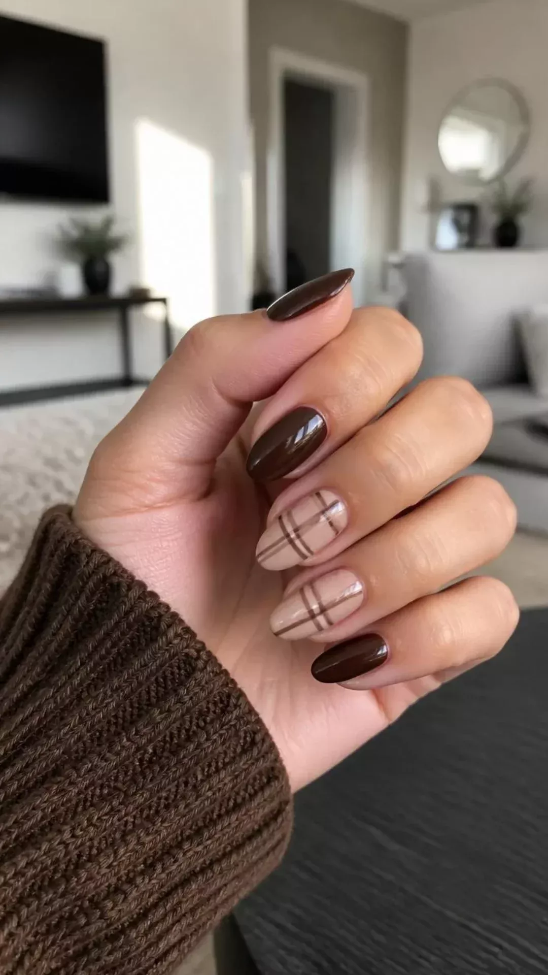 Close-up of a hand with brown and plaid pattern oval nails, wearing a brown knitted sweater.