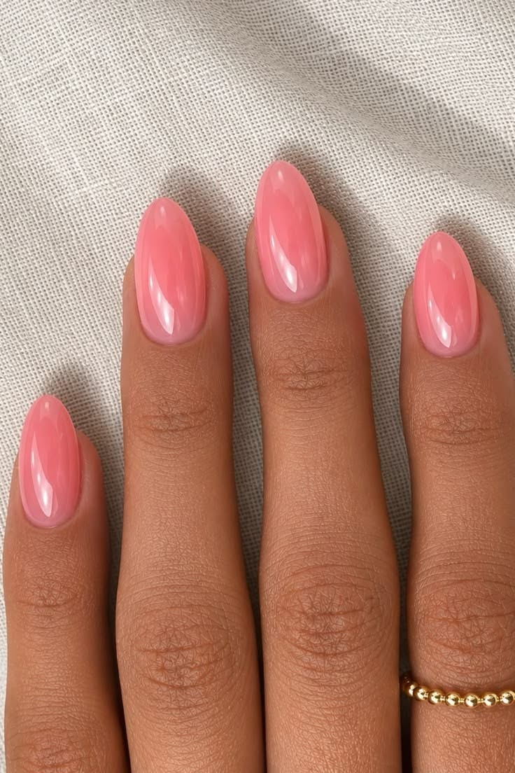 A hand with oval-shaped nails painted in a sheer, light pink polish. A gold beaded ring is on one finger.