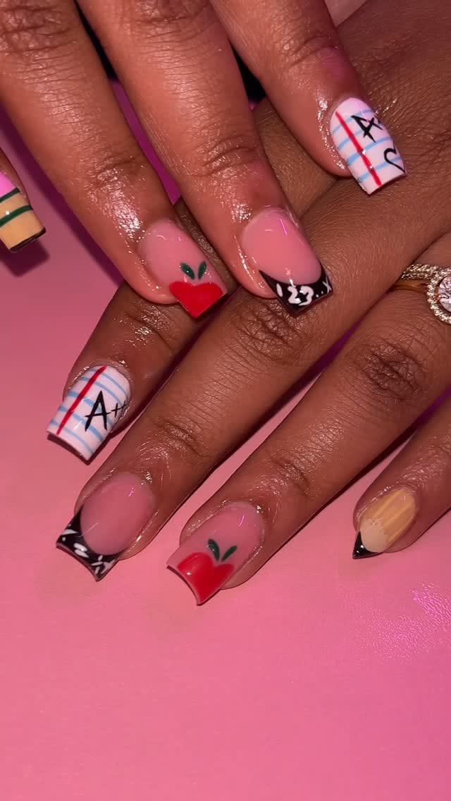 Close-up of a person's hands with square-tipped nails featuring various school-themed designs on a pink background.
