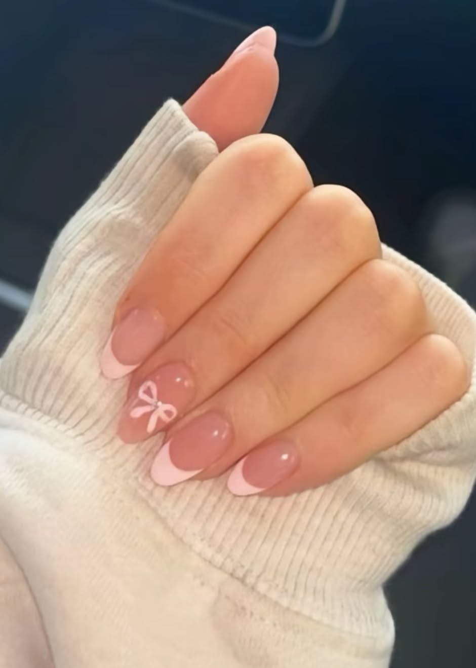 Close-up of a hand with almond-shaped nails, featuring a pink-tipped French manicure with a white bow design on the ring…