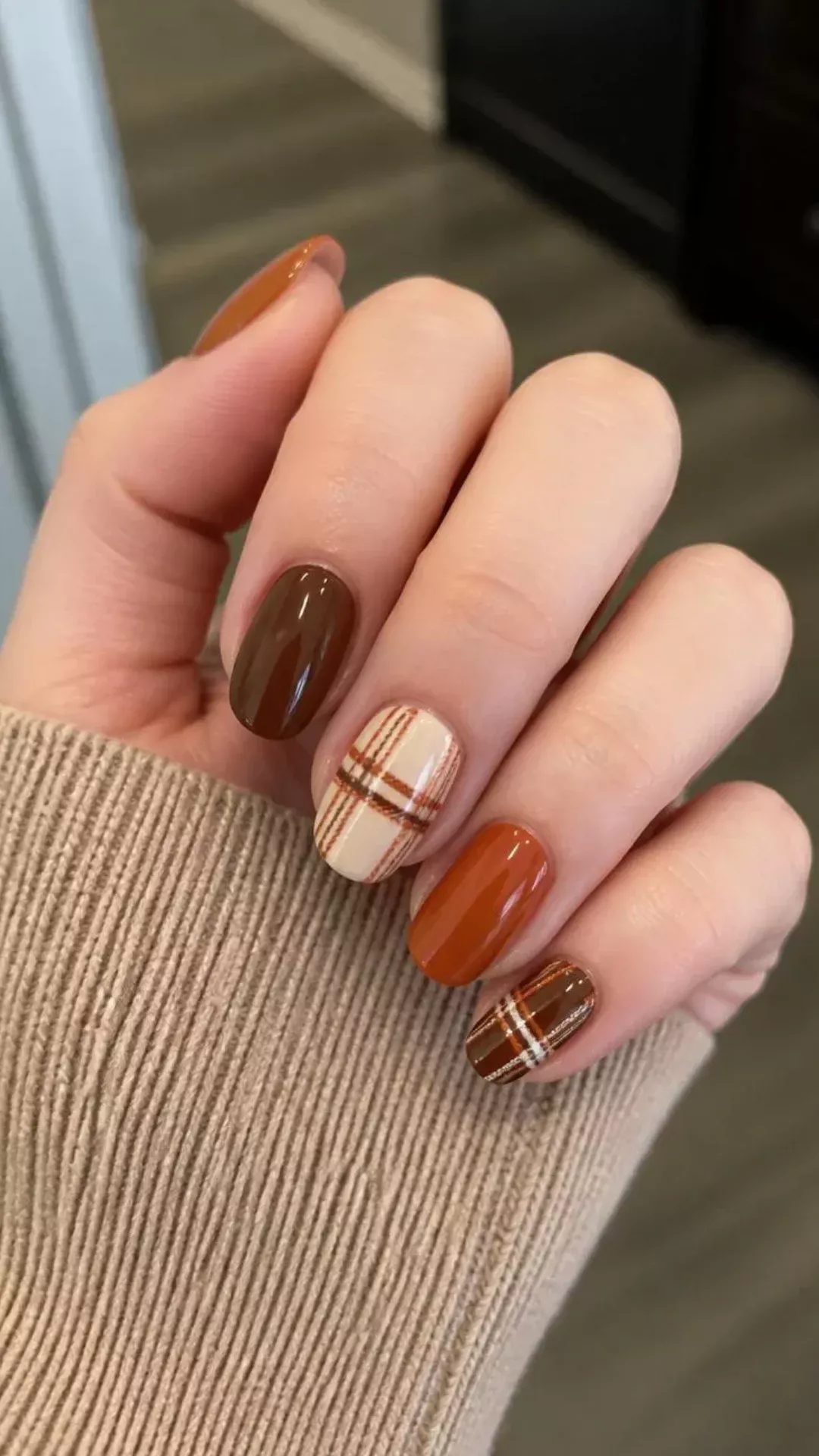 A hand with several fingernails painted with a plaid pattern in brown, orange, and cream, and other nails painted solid…