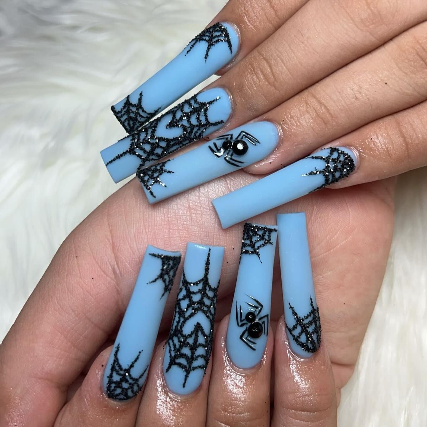 Long, square light blue nails with black glitter spiderweb designs and raised black spider accents.