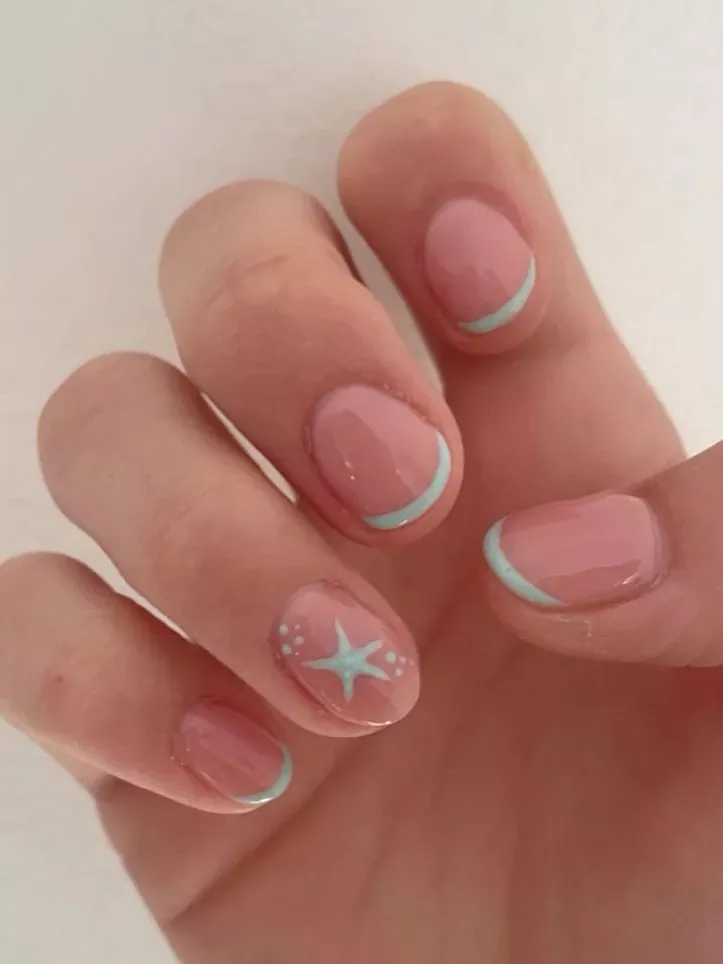 Close-up of a hand with short, pink manicured nails featuring light blue French tips and a small starfish design on one nail.