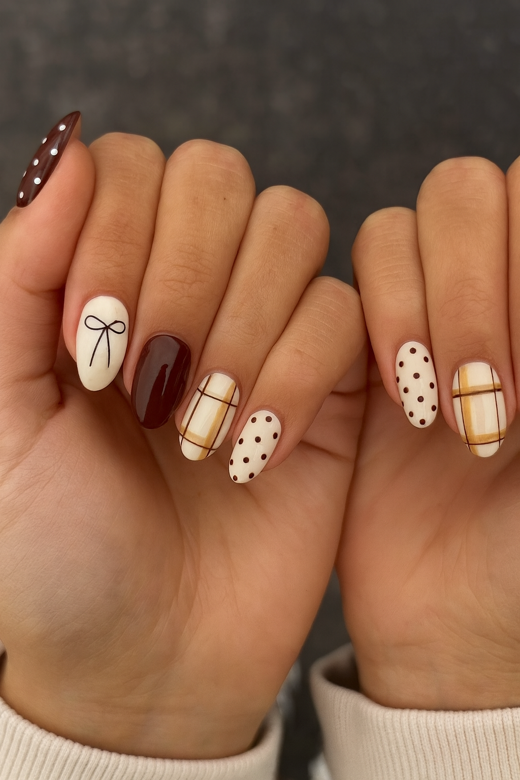 Hands with short, rounded nails painted with brown and cream patterns, including polka dots, plaid, and a bow design.