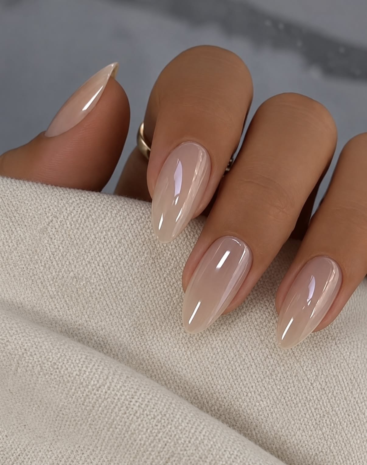 Hand with almond-shaped nails painted in a glossy, milky nude color, resting on a light-colored fabric.
