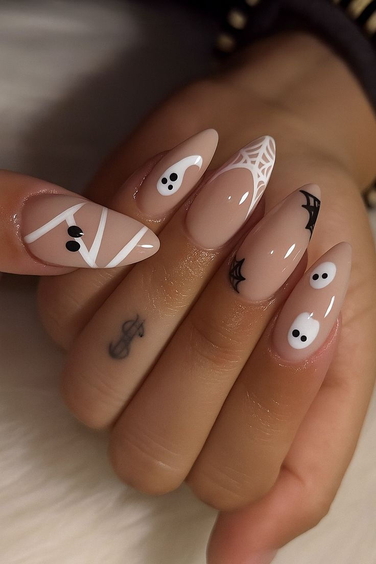 Almond-shaped nails with a neutral base feature different Halloween designs: mummies, ghosts, spiderweb tips, and a small…