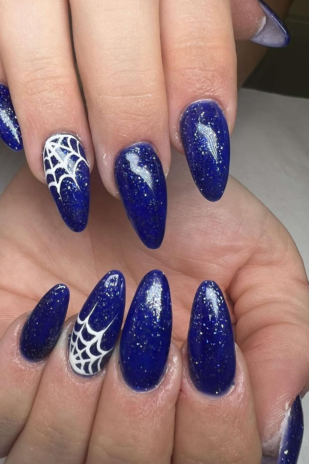 Close-up of a hand with dark blue almond-shaped nails, two of which feature white spiderweb designs.
