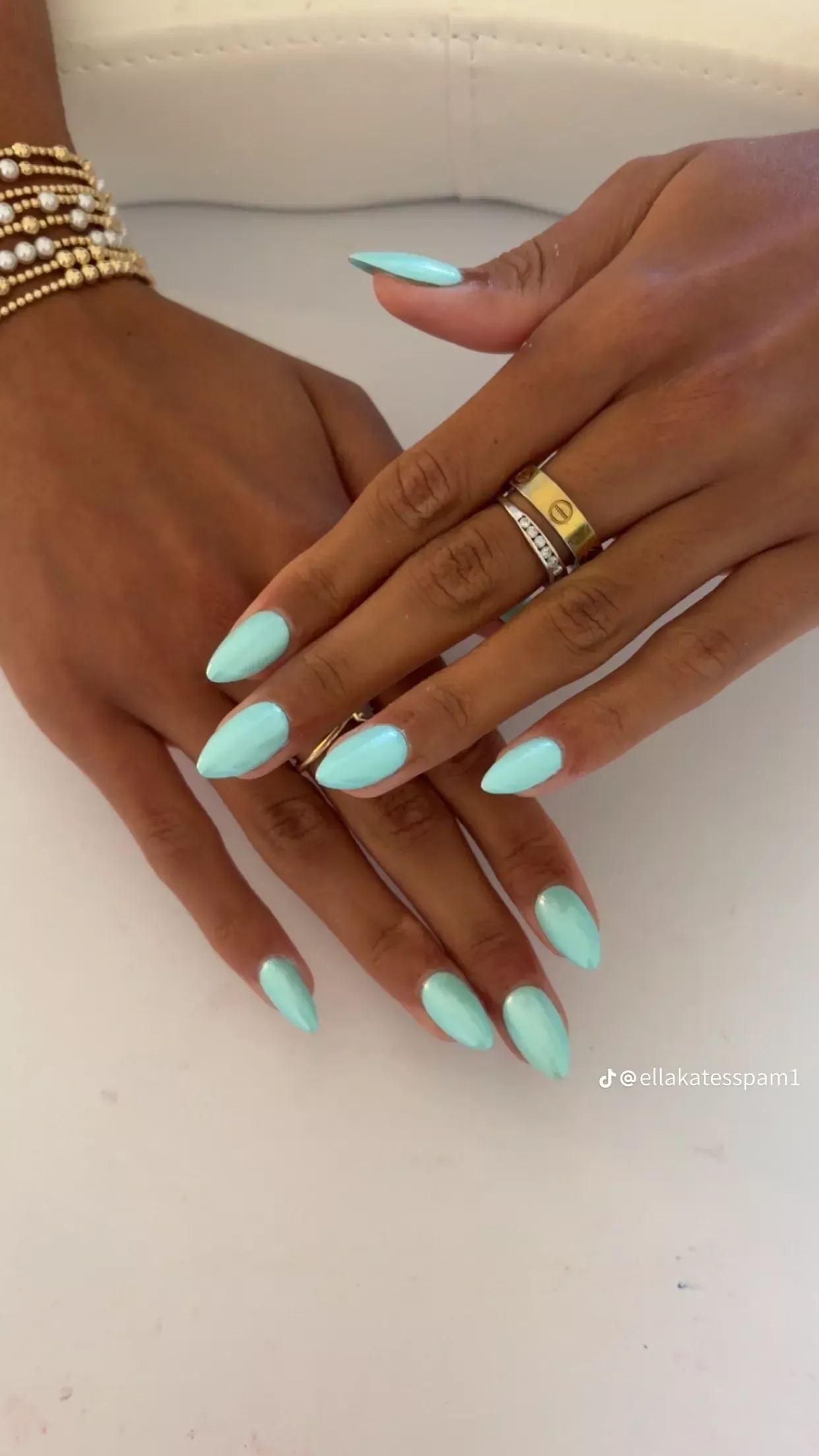 Close-up of hands with light aqua almond-shaped nails, wearing gold rings and a layered gold bracelet.