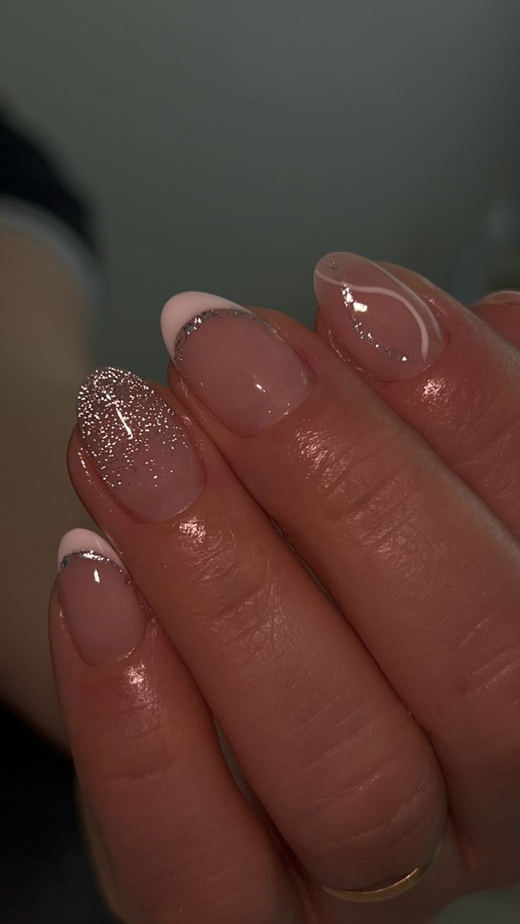 Close-up of a hand with short, oval nails, featuring a clear base, light pink tips, and a fine line of silver glitter just…