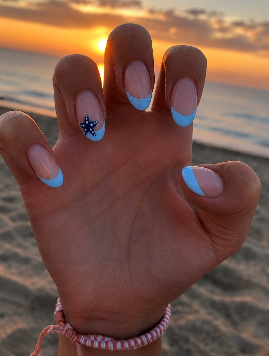 Hand with short nails in blue French manicure with one starfish accent nail, against a sunset beach backdrop.
