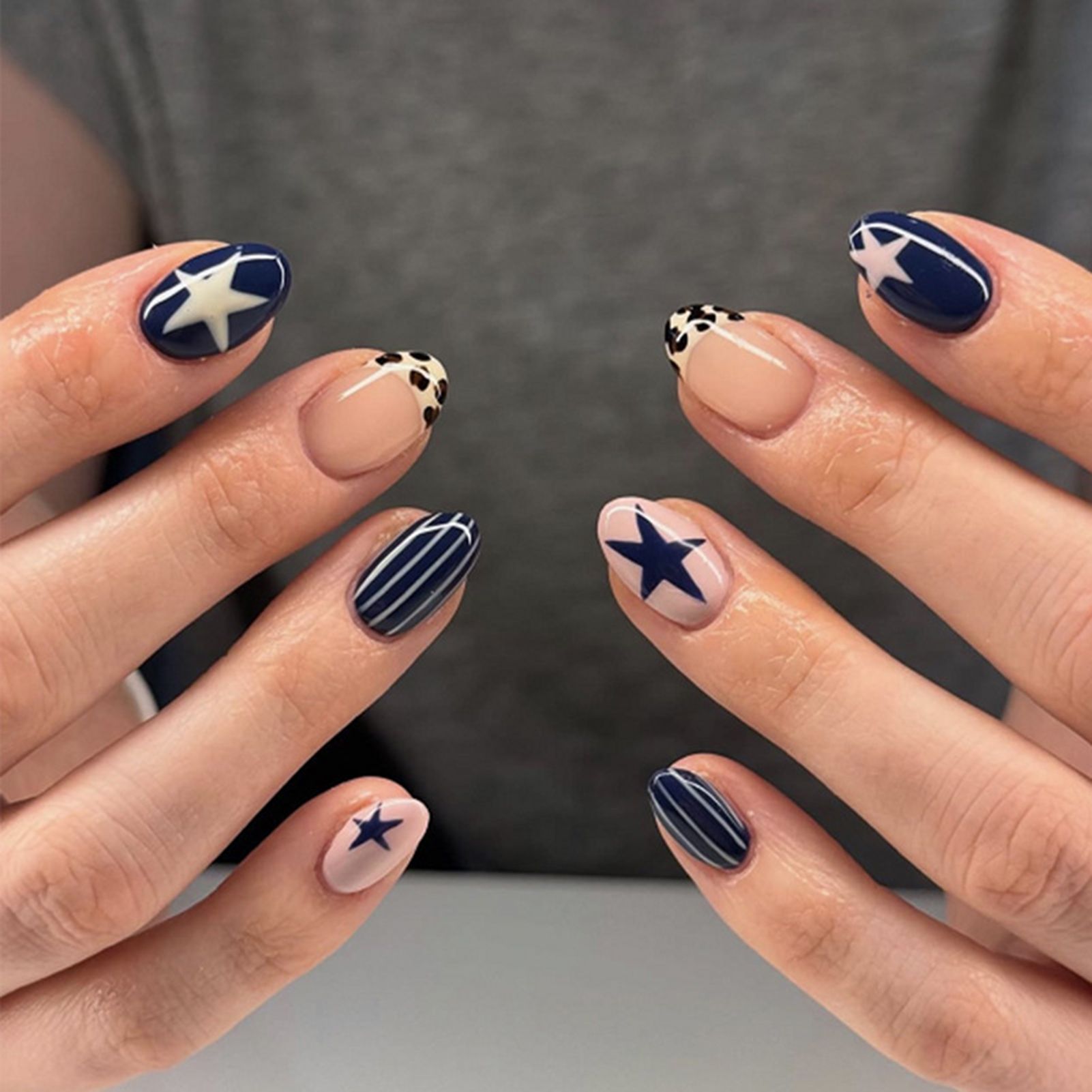 Close-up of hands with short oval nails, featuring navy blue and light pink polish with star, stripe, and leopard print…