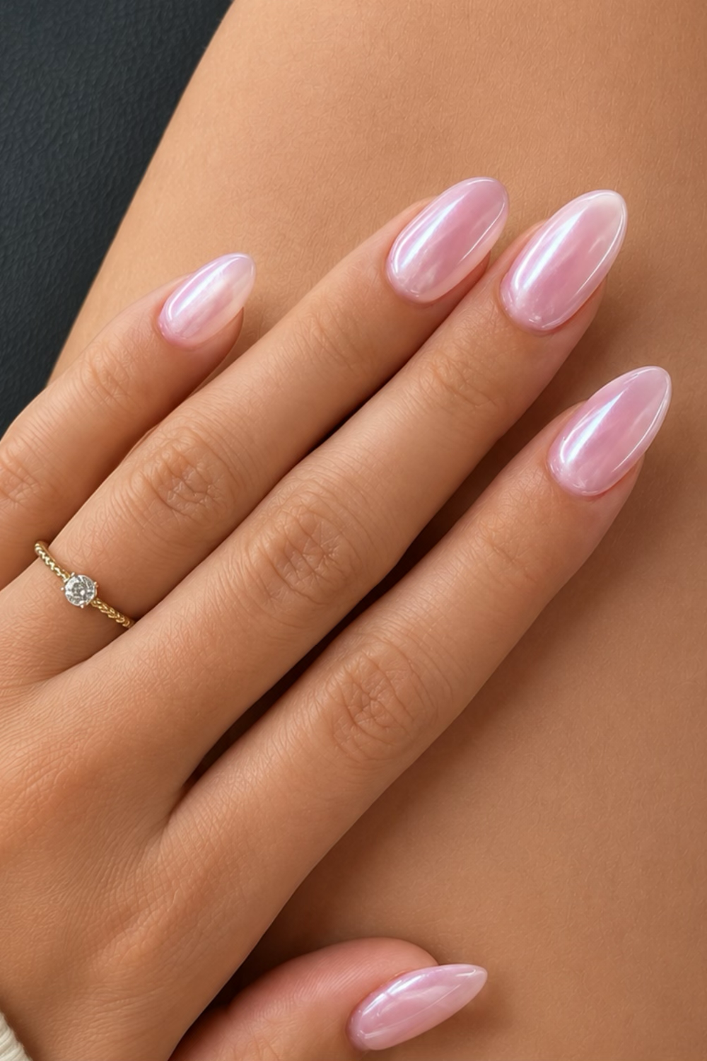 A hand with oval-shaped, iridescent pink nails, wearing a gold ring with a single clear gem on the ring finger.