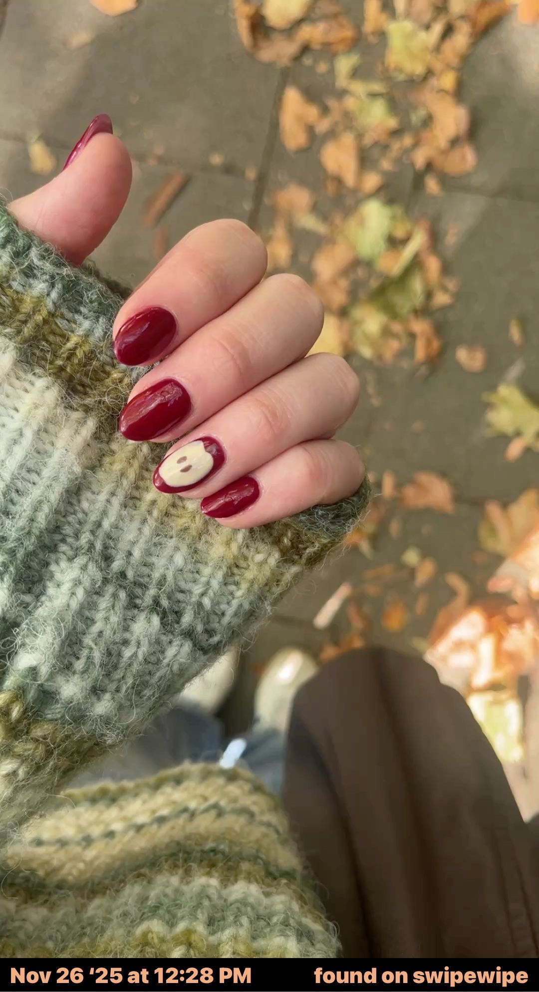 Hand with red manicured nails, one nail featuring a light-colored apple design, against a blurred background of fallen…