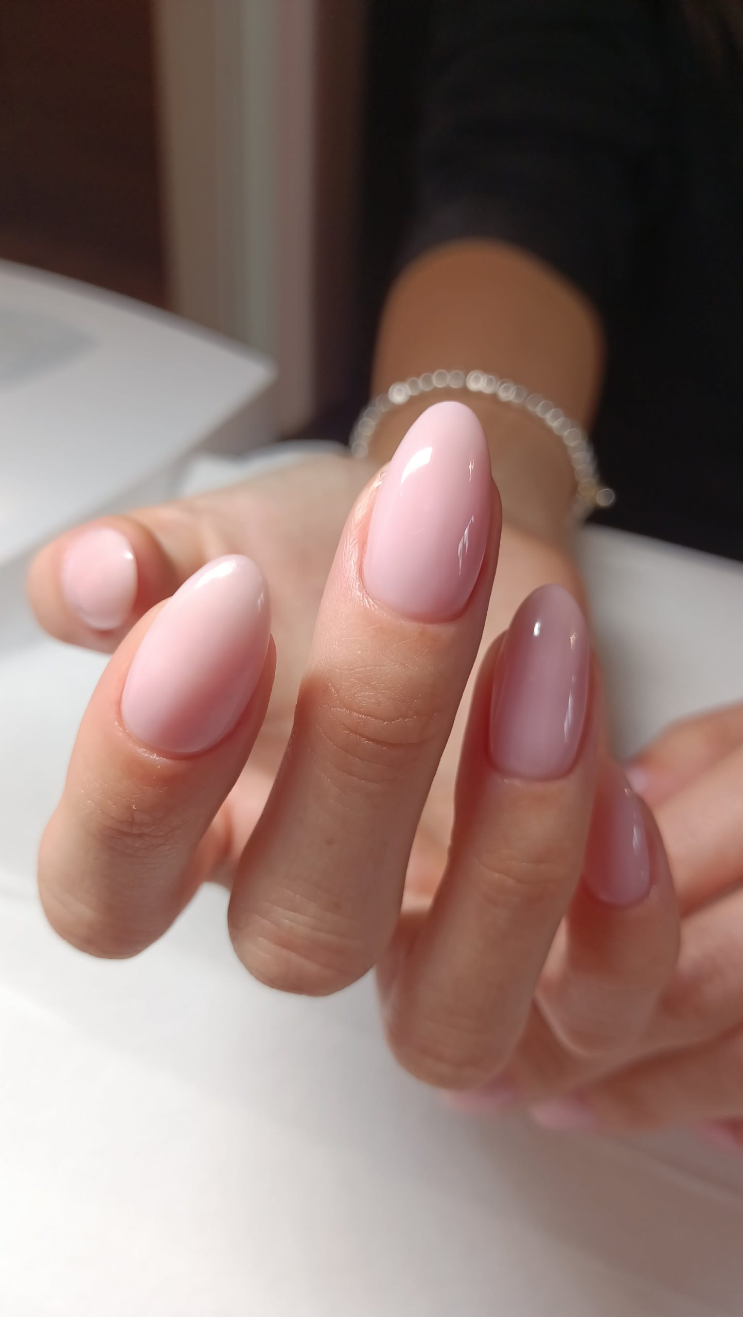 Close-up of a person's hand with light pink, oval-shaped gel nails. A silver chain bracelet is visible on the wrist.