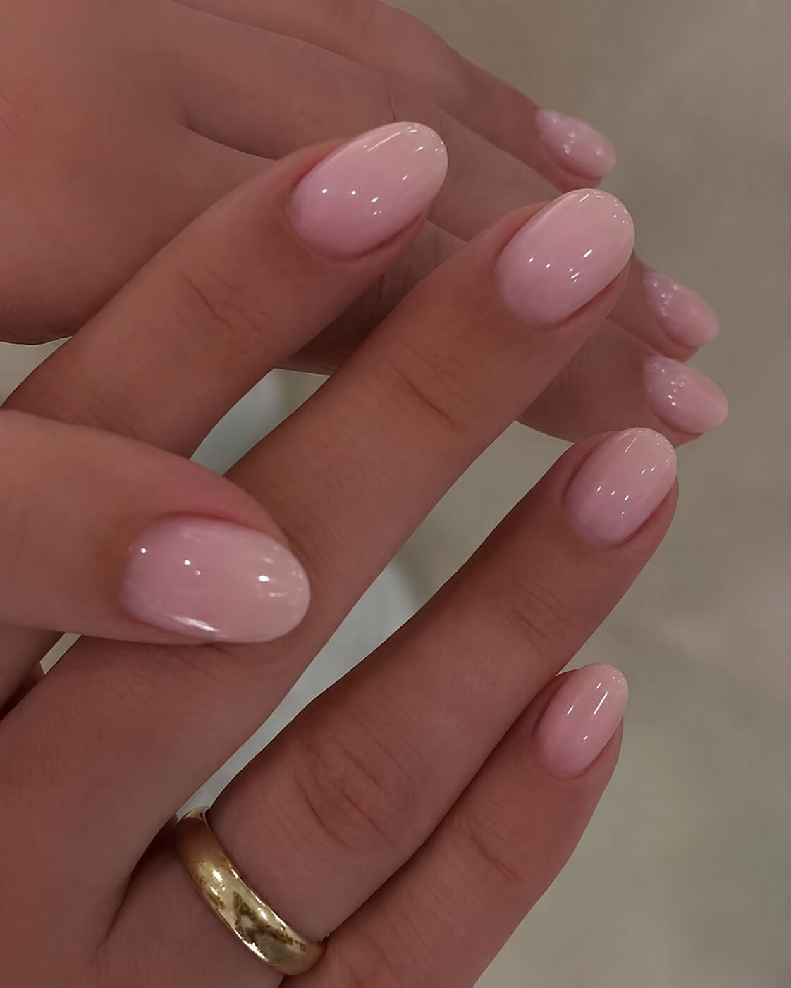 Hands with short, oval-shaped nails painted in a glossy, light pink polish. A gold ring is on one finger.