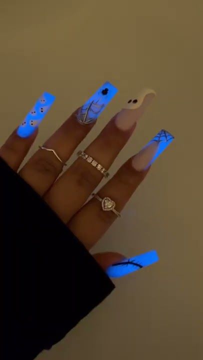 A hand with long, coffin-shaped nails featuring glowing blue Halloween designs like spiderwebs and ghosts.