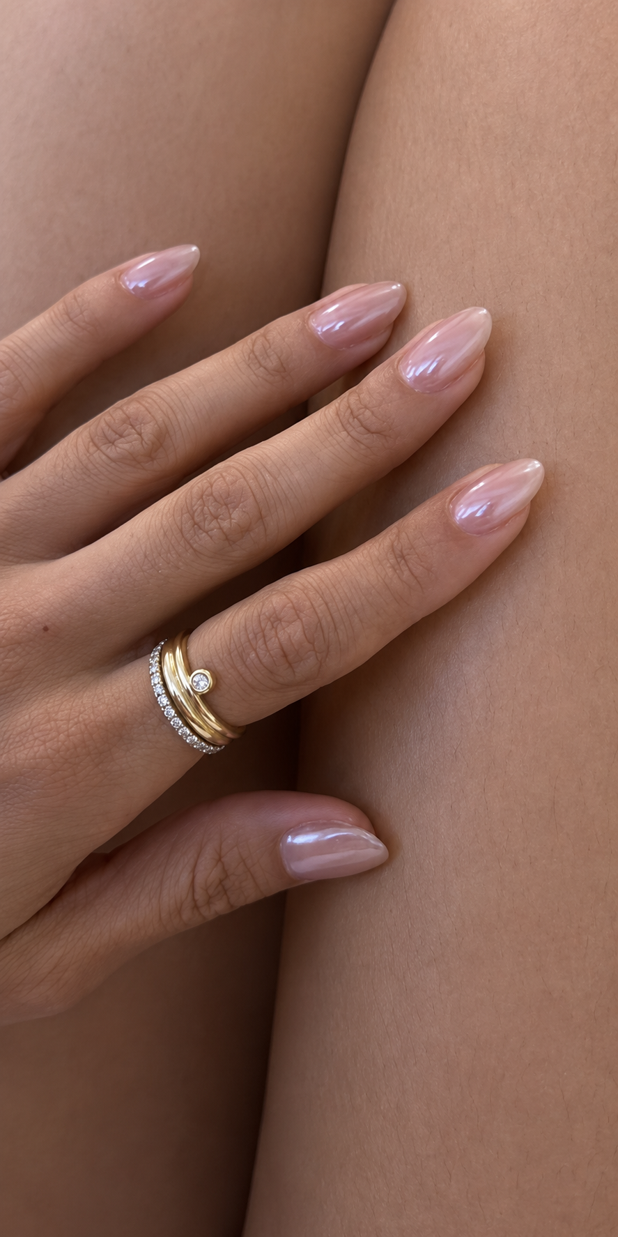 A hand with almond-shaped nails coated in a subtle, shimmery, natural chrome polish, wearing gold rings.