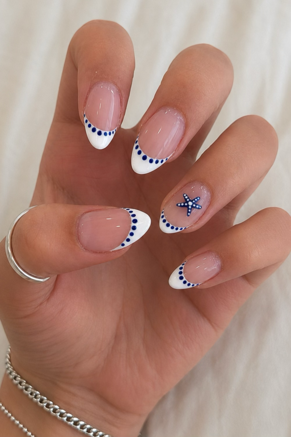 Hand with short, oval-shaped nails featuring white French tips with navy blue dots along the curve, and a small starfish…