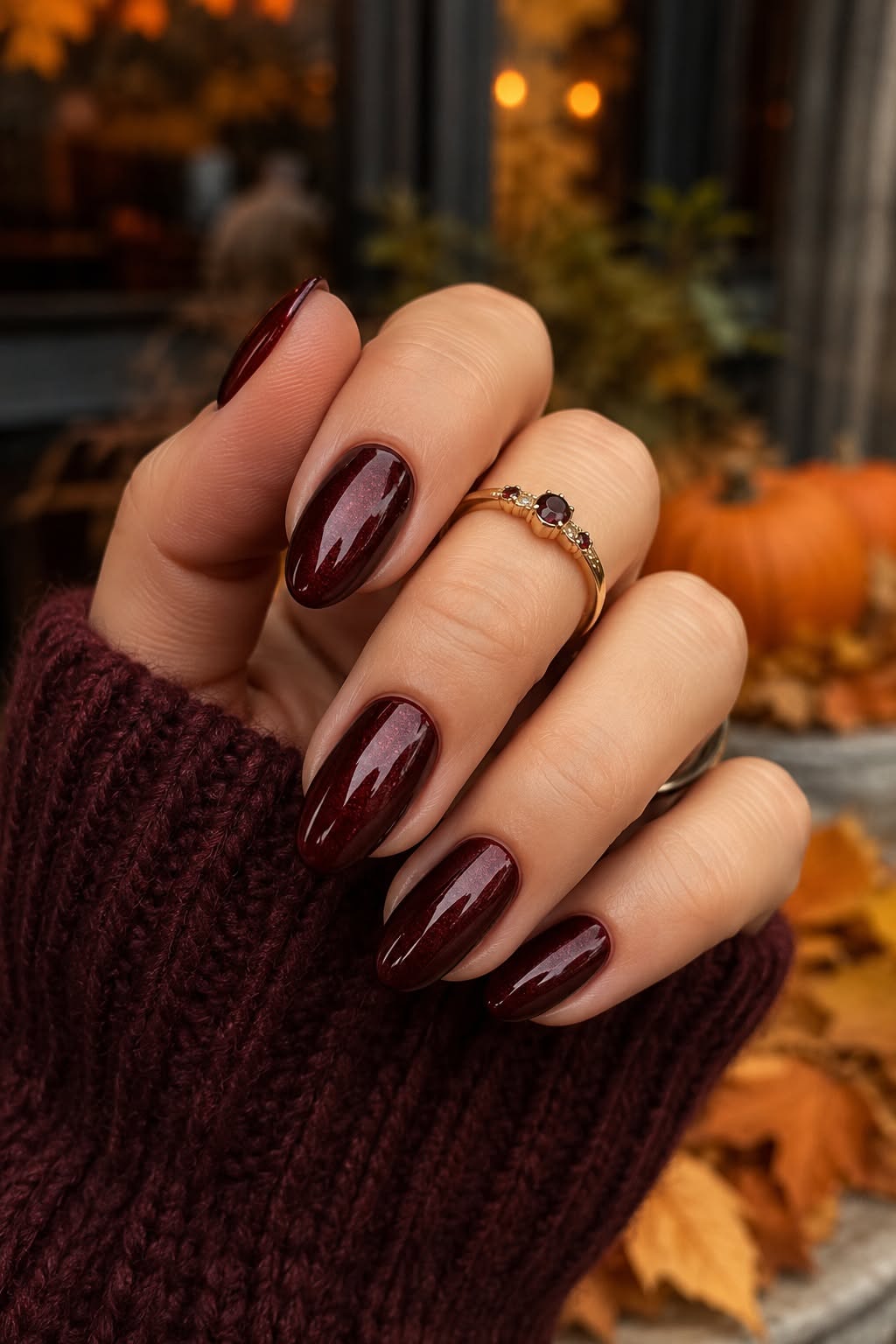 Hand with dark red oval nails and a gold ring, wearing a maroon sweater, against a blurred background of autumn leaves and a…