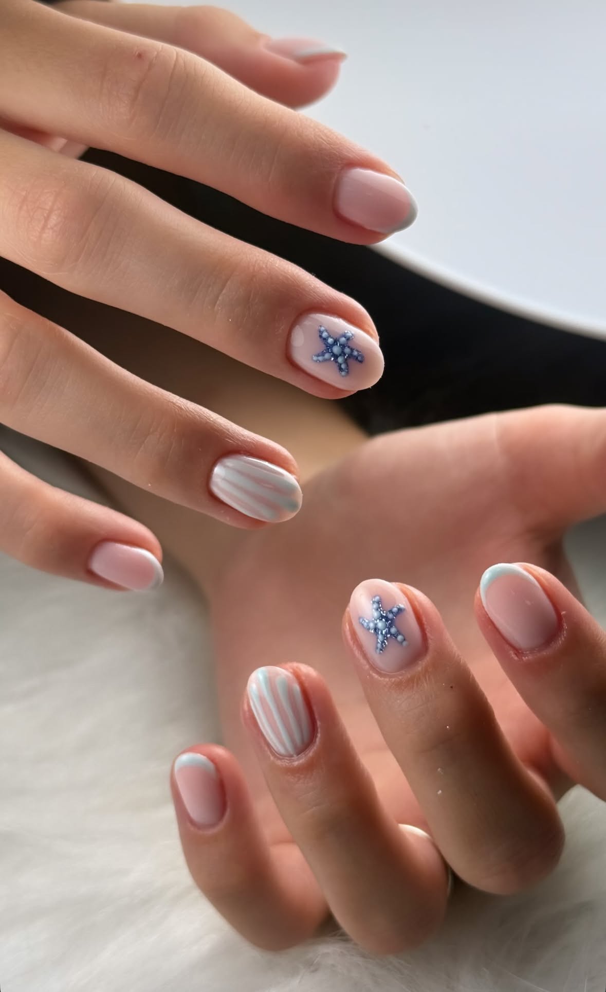 Close-up of hands with short nails featuring blue starfish and white striped designs, along with light blue french tips.