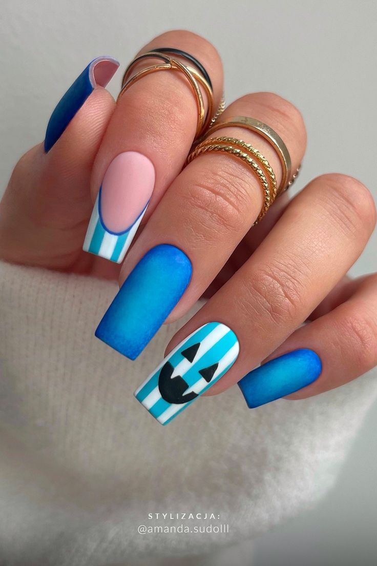 Close-up of a hand with long blue, white, and pink square-tipped nails, some with stripes and a pumpkin face.