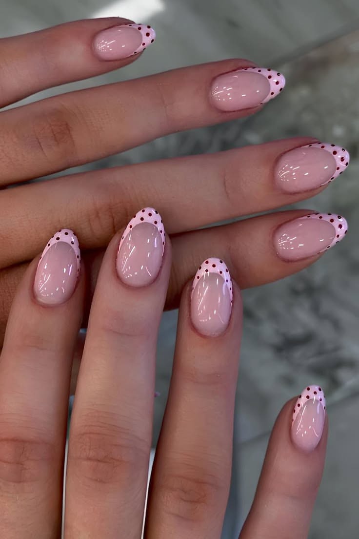 Close-up of a person's hands with short, oval-shaped nails featuring a clear base and pink polka-dotted French tips.