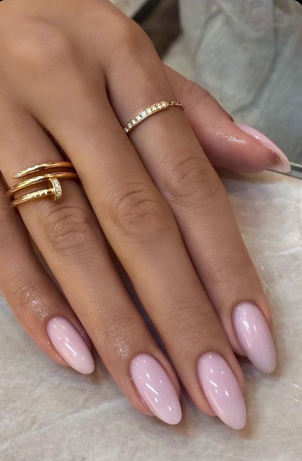 A hand with long, oval-shaped nails painted in a sheer milky pink polish, wearing two gold rings on different fingers.
