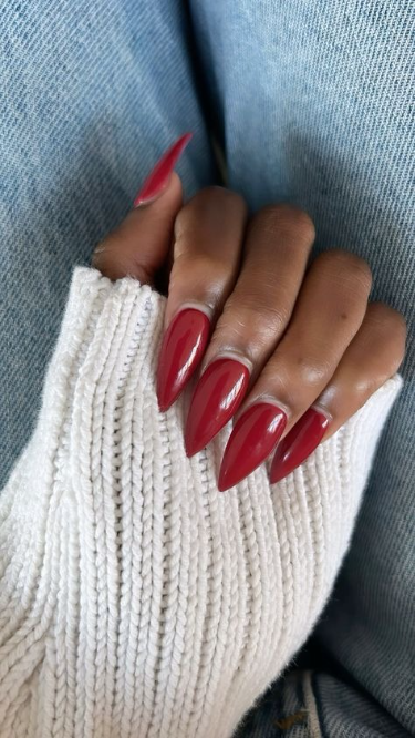 Hand with glossy red stiletto nails, wearing a white knitted sweater cuff against blue denim.