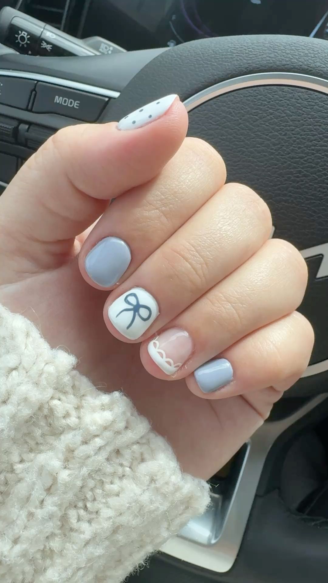A person's hand with short nails in light blue and white. One nail has a bow, another has a scallop design.