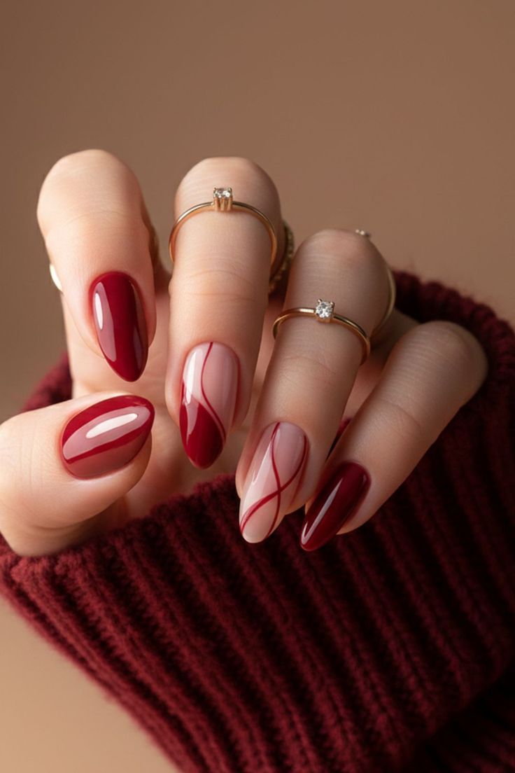 A hand with oval-shaped nails, some solid red, others clear with red swirls and lines. The hand wears gold rings.
