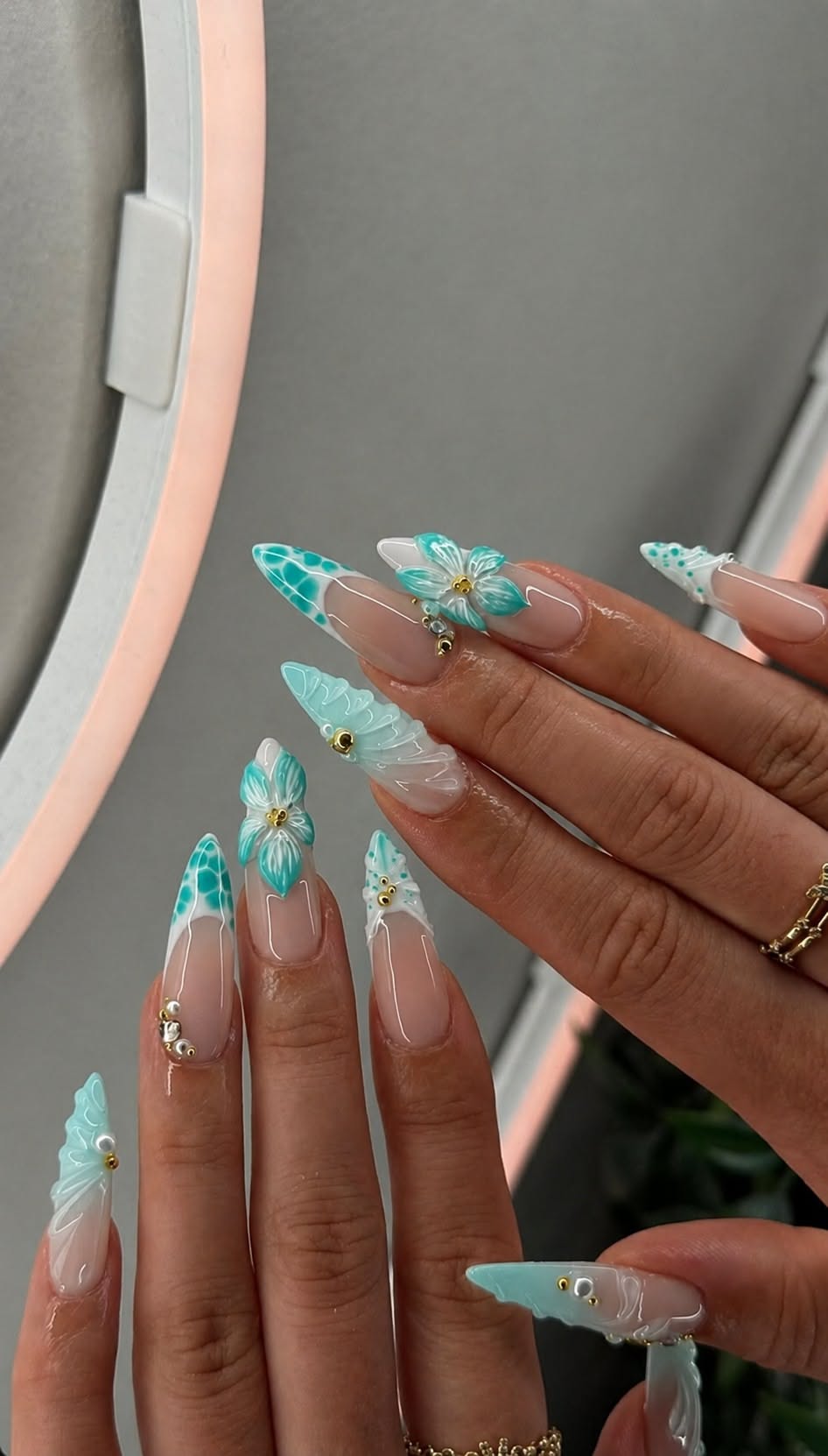 Pointed almond-shaped nails with clear bases, white French tips, aqua wave patterns, 3D flowers, and gold bead accents.