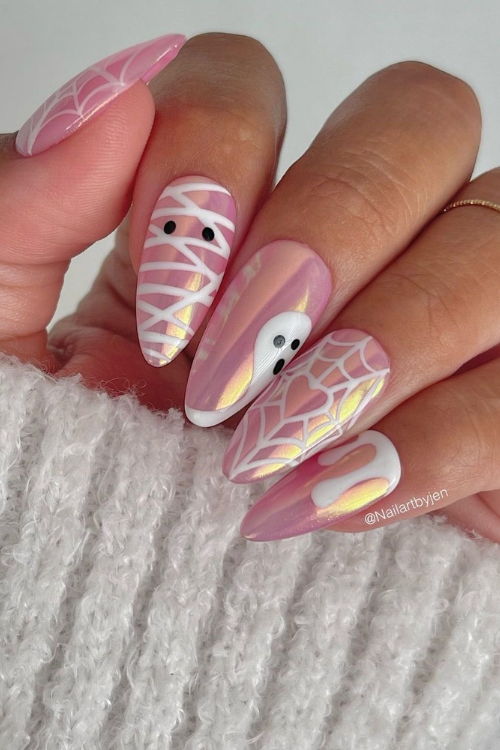 Close-up of a hand with iridescent pink Halloween-themed nails, featuring designs of a ghost, a mummy, and spiderwebs.