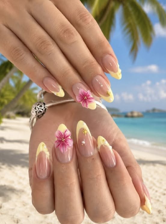 Close-up of a person's hands with long oval nails featuring yellow French tips, 3D pink flowers, and tiny gold and white…