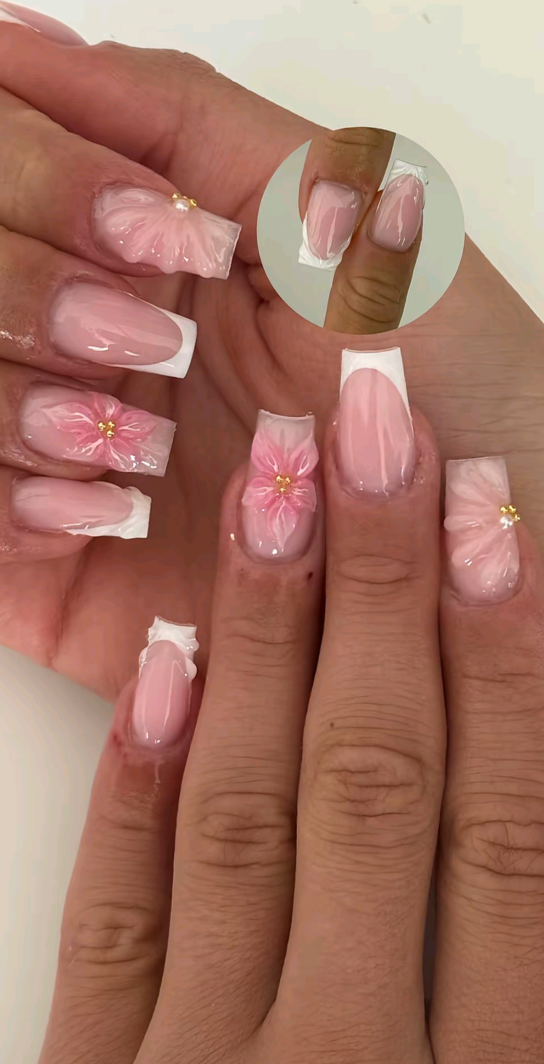 Close-up of a hand with pink and white french manicure style nails, some adorned with raised pink flower designs and small…