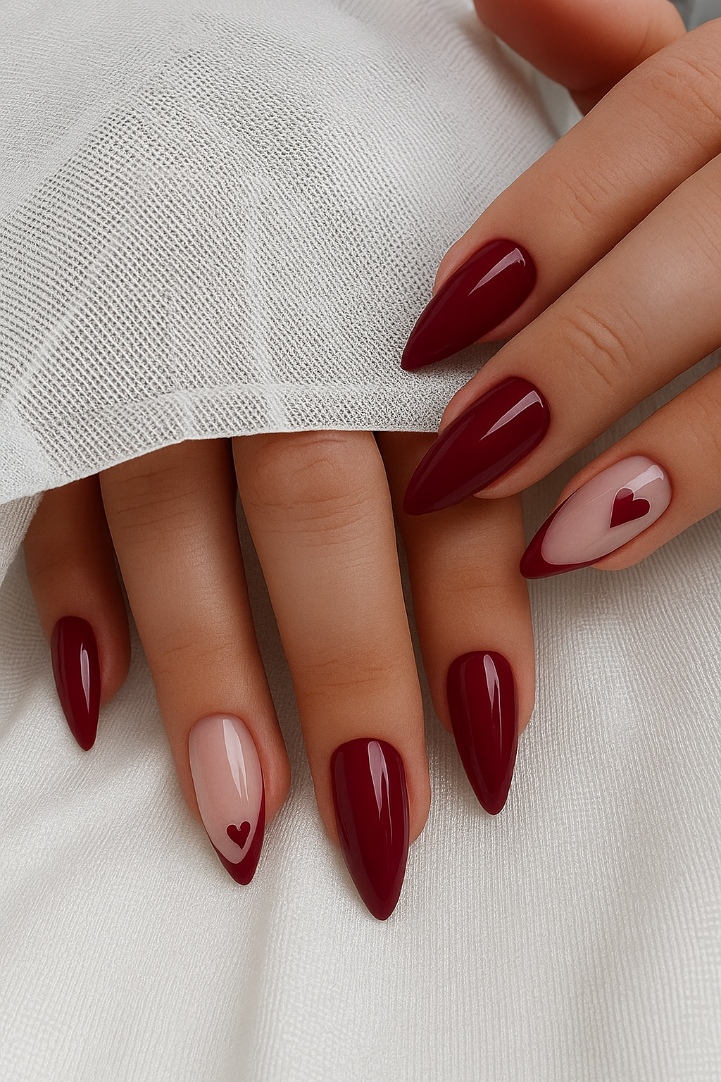 Burgundy almond-shaped nails with a glossy finish. Two accent nails feature a light pink base with a small red heart design…
